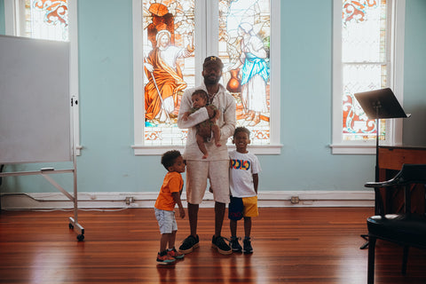 Tio Fallen stands in front of a stained glass window, wearing all white. His two older sons are on either side of him, and he holds his youngest son in his arms. 
